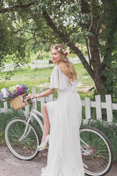 Beautiful smiling young bride in wedding dress riding bicycle — Stock Photo
