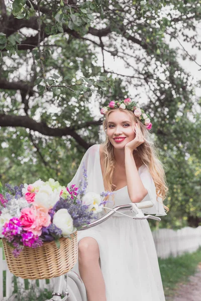 Bela jovem noiva andar de bicicleta com cesta de flores e sorrindo para a câmera — Fotografia de Stock