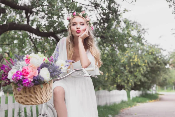 Beautiful young bride riding bicycle with flower basket and looking at camera — Stock Photo