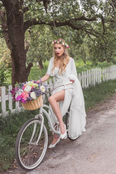 Beautiful shocked young blonde bride riding bicycle with flower basket — Stock Photo