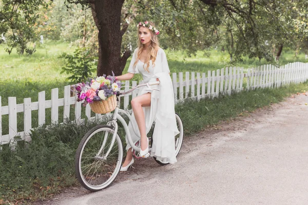 Beautiful shocked bride in wedding dress riding bicycle and looking away — Stock Photo