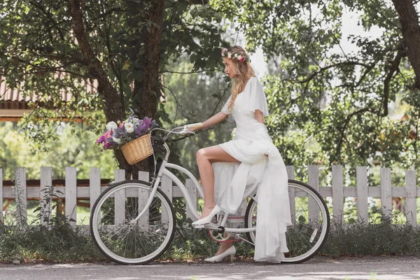 Vista lateral de la hermosa novia joven en vestido de novia a caballo bicicleta y mirando hacia otro lado - foto de stock