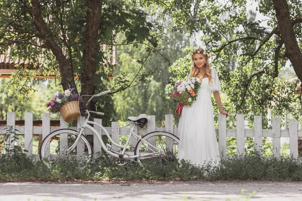 Bella giovane sposa che tiene bouquet e sorride alla macchina fotografica mentre in piedi vicino alla bicicletta alla recinzione — Foto stock