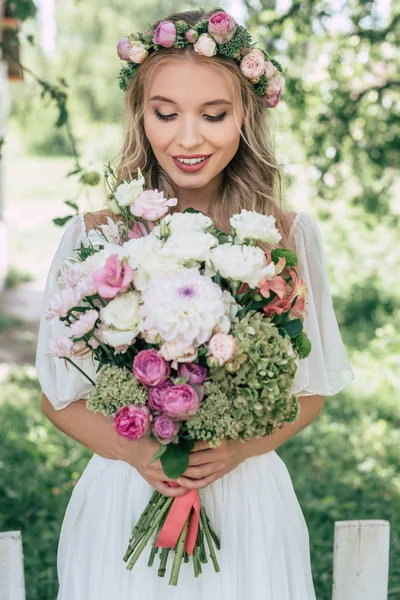 Attractive smiling bride in floral wreath holding beautiful bouquet of flowers outdoors — Stock Photo