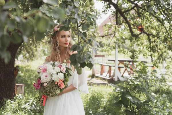 Schöne nachdenkliche Braut hält Brautstrauß in der Hand und schaut im Garten weg — Stockfoto