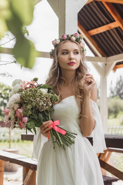 Belle jeune mariée réfléchie tenant bouquet de fleurs et regardant loin — Photo de stock