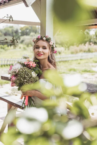 Foco seletivo de bela noiva jovem em grinalda floral segurando buquê de casamento e sorrindo para a câmera — Fotografia de Stock