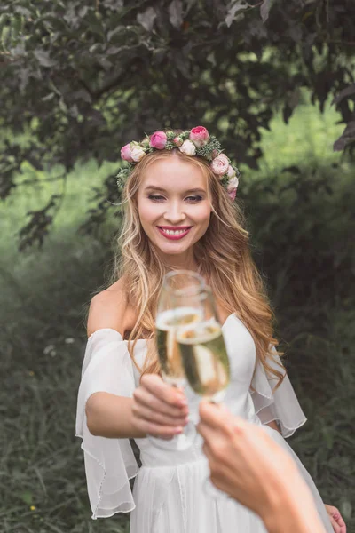 Beautiful smiling young bride clinking glasses of champagne with someone in park — Stock Photo