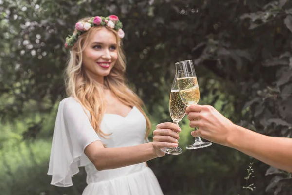 Cropped shot of happy young bride clinking glasses of champagne with someone in park — Stock Photo
