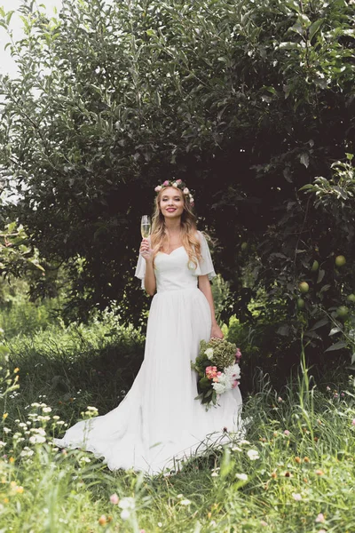 Hermosa novia joven celebración de ramo de boda y copa de champán, sonriendo a la cámara en el jardín - foto de stock