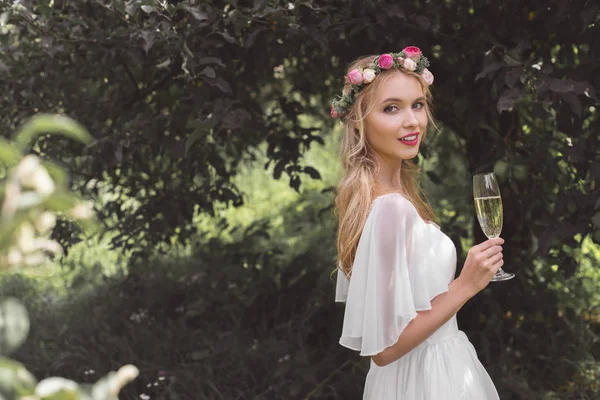 Beautiful young bride holding glass of champagne and smiling at camera outdoors — Stock Photo