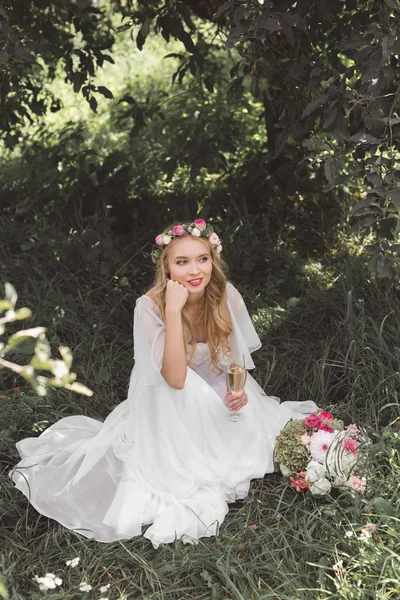 Vista ad alto angolo di bella giovane sposa sorridente seduta sull'erba e con in mano un bicchiere di champagne — Foto stock