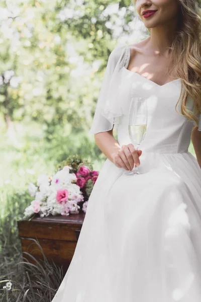 Cropped shot of smiling young bride holding glass of champagne and sitting on vintage chest — Stock Photo