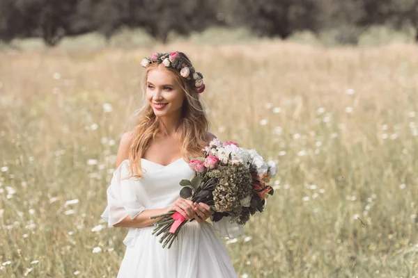 Beautiful smiling young bride holding wedding bouquet and looking away outdoors — Stock Photo