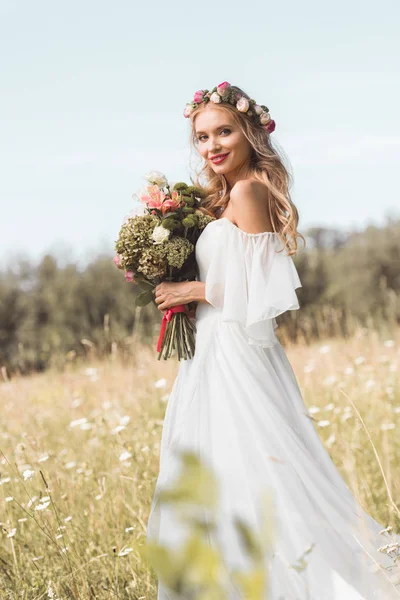 Bela noiva jovem em vestido de noiva e coroa floral segurando buquê e sorrindo para a câmera — Fotografia de Stock