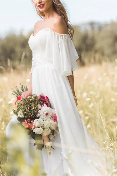 Plan recadré de jeune mariée souriante en robe de mariée tenant bouquet de fleurs à l'extérieur — Photo de stock