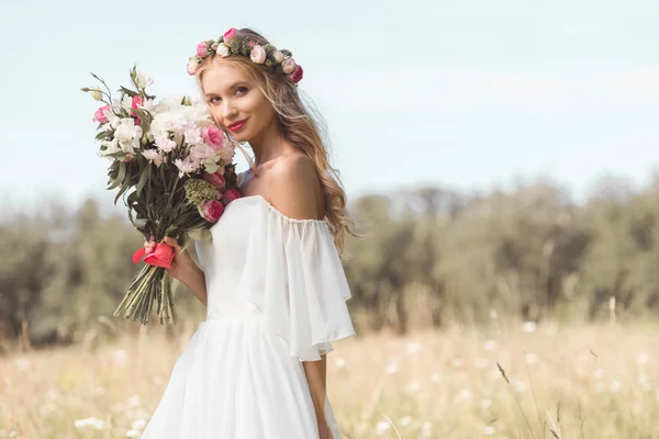 Bella giovane sposa che tiene bouquet da sposa e sorride alla macchina fotografica all'aperto — Foto stock