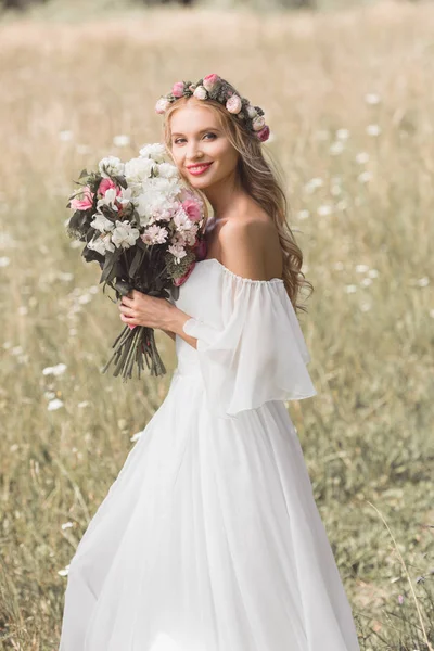 Hermosa joven novia en floral corona celebración de la boda ramo y sonriendo a la cámara al aire libre - foto de stock