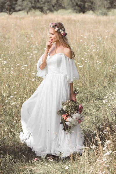Bela noiva pensativa em vestido de noiva e coroa floral segurando buquê de flores no campo — Fotografia de Stock