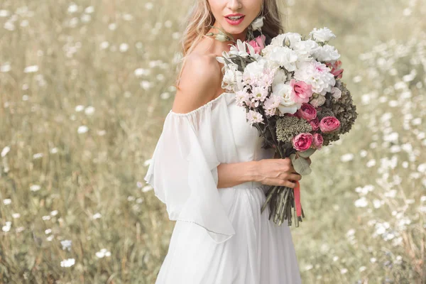 Cropped shot of young blonde bride in wedding dress holding beautiful bouquet of flowers outdoors — Stock Photo