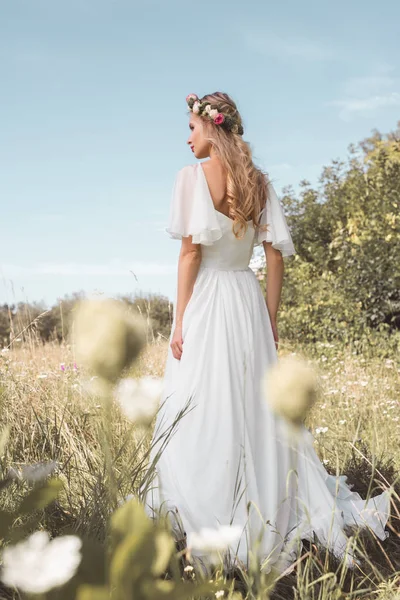 Back view of beautiful young bride in wedding dress and floral wreath walking on field — Stock Photo