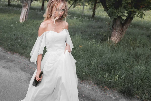 Vista ad alto angolo di sconvolto giovane sposa in abito da sposa in possesso di bottiglia di vino e camminare nel parco — Foto stock