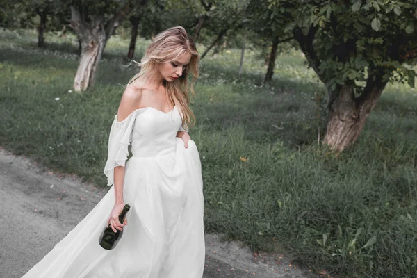 Upset young bride in wedding dress holding bottle of wine and walking in park — Stock Photo