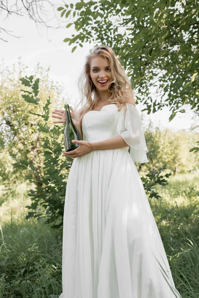 Low angle view of young bride holding bottle of wine and smiling at camera — Stock Photo