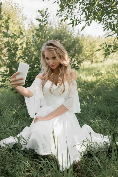 Mujer rubia joven en vestido de novia sentado en la hierba y tomando selfie con teléfono inteligente - foto de stock