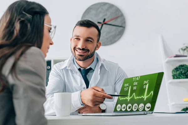 Sonriente doctor apuntando en portátil con aparato de pulso al paciente en la clínica - foto de stock