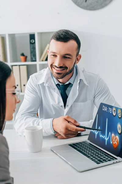 Médecin souriant pointant sur ordinateur portable avec application médicale au patient dans la clinique — Photo de stock