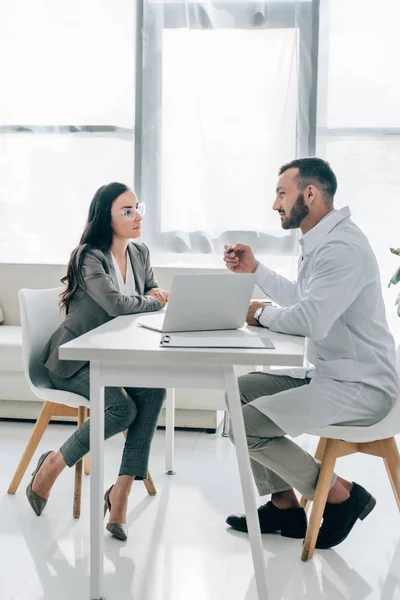 Seitenansicht von Patient und Arzt im Gespräch in der Klinik mit Laptop auf dem Tisch — Stockfoto