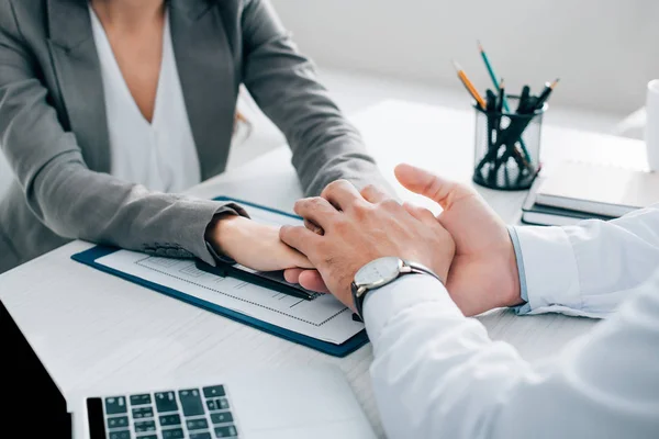 Image recadrée du patient et du médecin généraliste tenant la main au-dessus du formulaire de réclamation d'assurance en clinique — Photo de stock