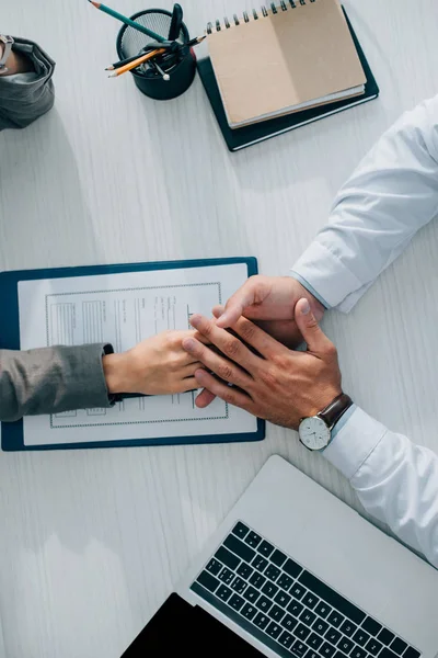 Cropped image of patient and doctor holding hands above insurance claim form in clinic — Stock Photo