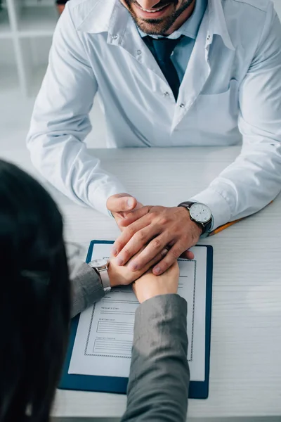 Обрезанное изображение пациента и врача, держащихся за руки над страховой формой в клинике — стоковое фото