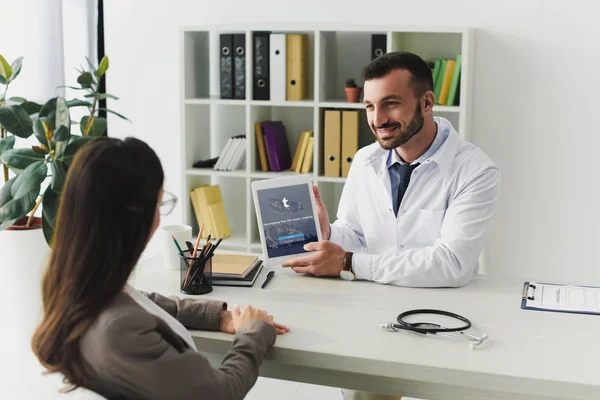 Médico sonriente mostrando tableta con página de tumblr cargada al paciente en la clínica - foto de stock