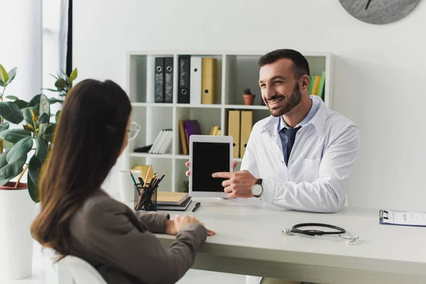 Médico sonriente apuntando en la tableta con pantalla en blanco al paciente en la clínica - foto de stock
