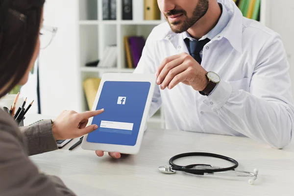 Cropped image of doctor showing patient tablet with loaded facebook page in clinic — Stock Photo