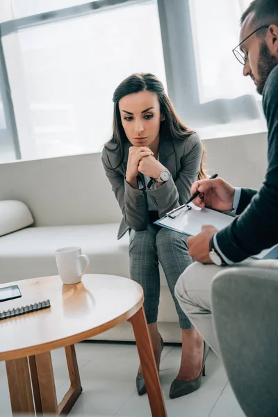 Paciente descansando la barbilla en las manos y psicólogo tomando notas al portapapeles en el consultorio de médicos - foto de stock