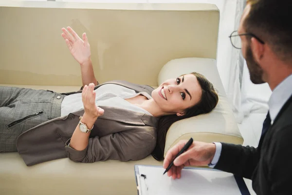 Happy patient lying on sofa and talking to psychologist in doctors office — Stock Photo