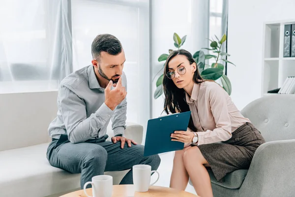 Psicólogo hablando con el paciente masculino y mostrando portapapeles en el consultorio - foto de stock