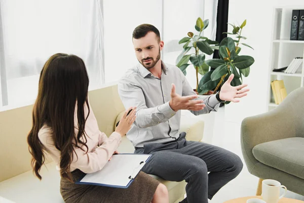 Professional psychiatrist with clipboard talking with patient in bad mood — Stock Photo