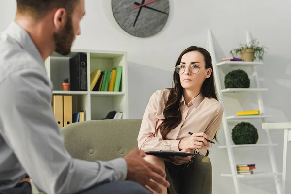 Psiquiatra profesional con portapapeles hablando con el paciente masculino en el consultorio - foto de stock