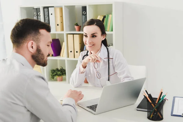 Sonriente médico y hombre discutiendo el seguro de salud y mirando en la pantalla del ordenador portátil en la clínica - foto de stock