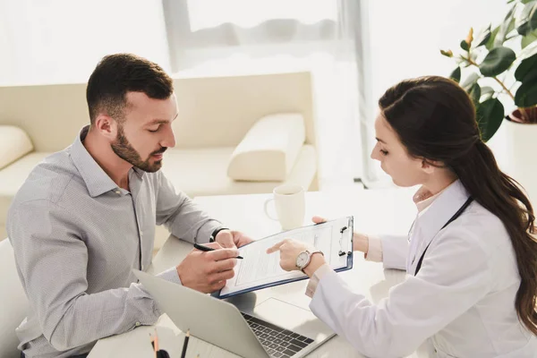 Mann unterschreibt Versicherungsantragsformular, während Arzt im Büro mit Laptop darauf zeigt — Stockfoto