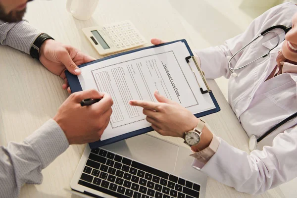 Visão cortada do cliente assinar formulário de pedido de seguro, enquanto o médico apontando para ele na clínica com laptop e calculadora — Fotografia de Stock