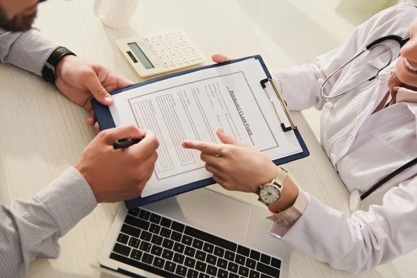 Vista ritagliata del cliente firma modulo di richiesta di assicurazione, mentre il medico che indica in clinica con computer portatile e calcolatrice — Foto stock