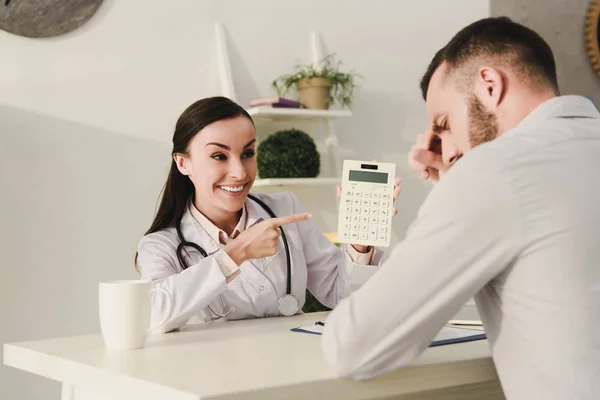 Cliente sonriente y médico profesional que cuenta las finanzas en la calculadora para el seguro de vida en la oficina - foto de stock