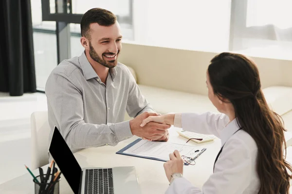 Cliente sonriente estrechando la mano con el médico en la oficina con el ordenador portátil y el formulario de reclamación de seguro - foto de stock