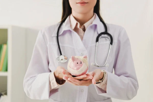 Cropped view of female doctor in white coat holding bandaged piggy bank — Stock Photo
